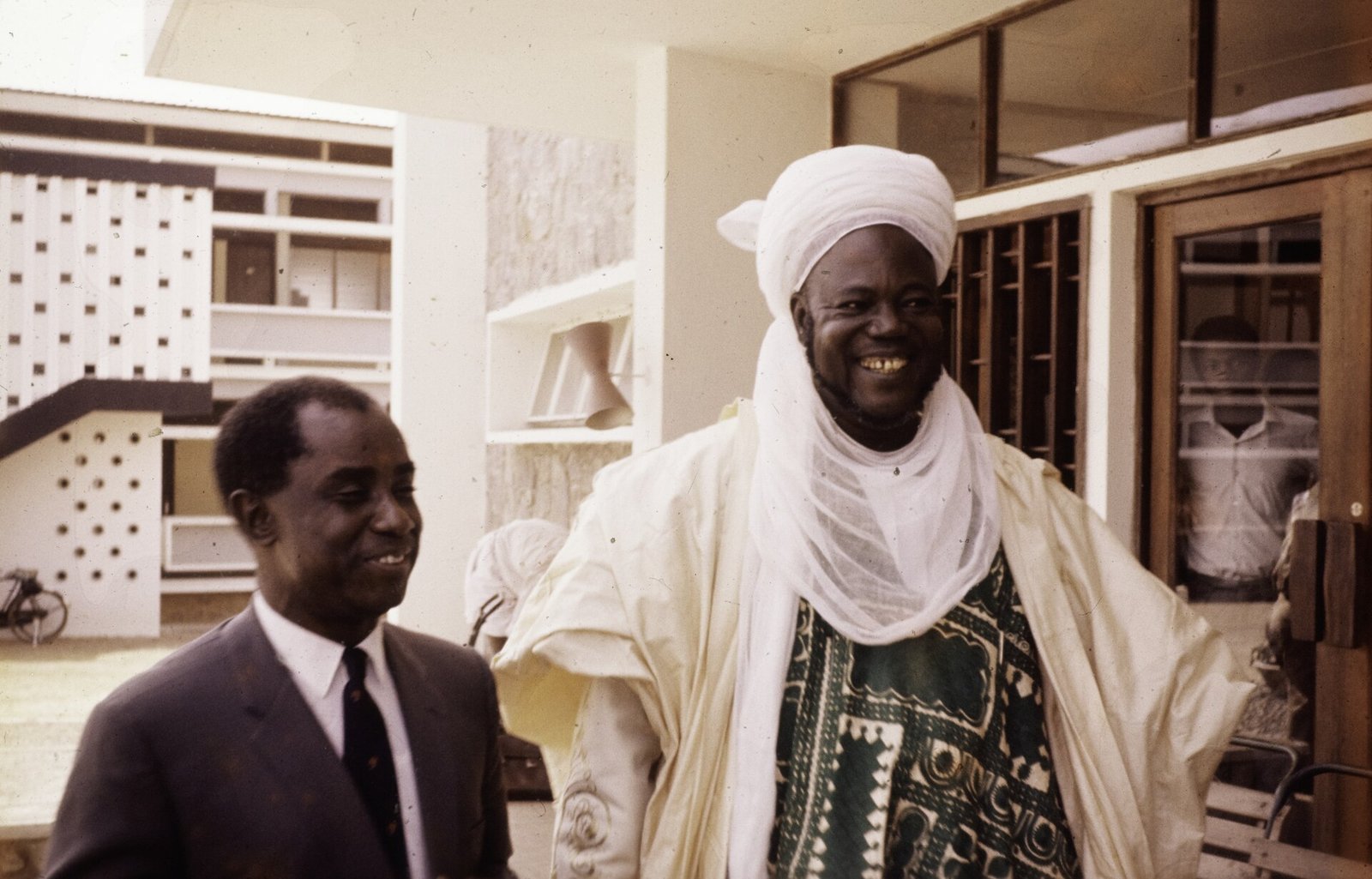 A smiling man in a white turban and a white robe over a green patterned gown stands beside a smiling man in a grey suit and tie, in front of a modern building with patterned concrete screens.