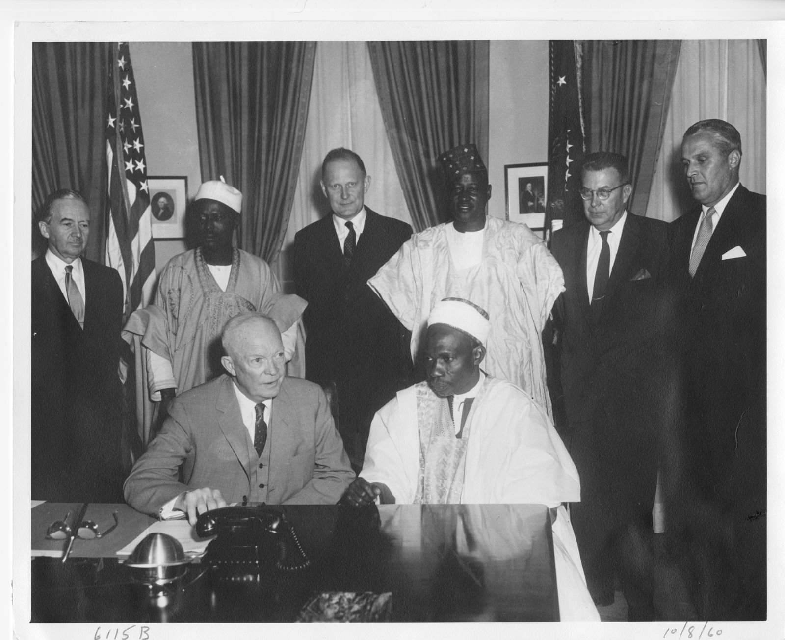 President Eisenhower seated at his desk beside a Nigerian man in a white robe and cap, with two Nigerians in robes and caps and four men in dark suits standing behind them, flags and framed portraits at the back.