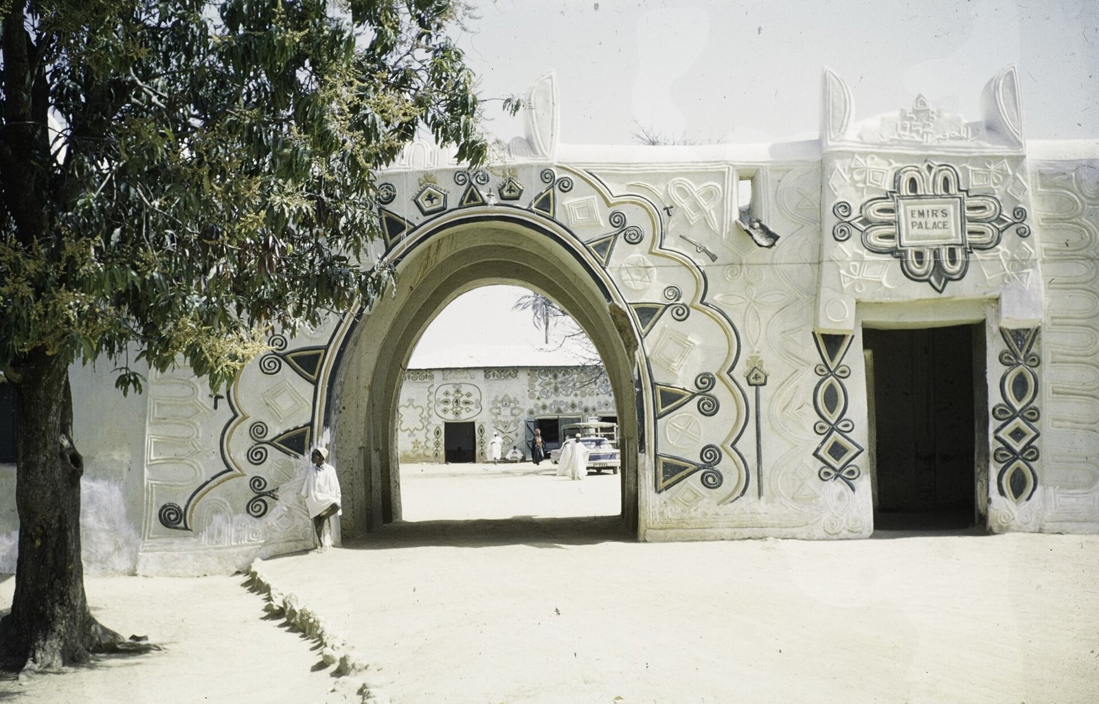 A whitewashed gateway with a pointed arch and raised geometric decoration, a plaque reading Emir's Palace, a man in white leaning against the wall in the shade of a tree, and a courtyard with a parked car beyond.