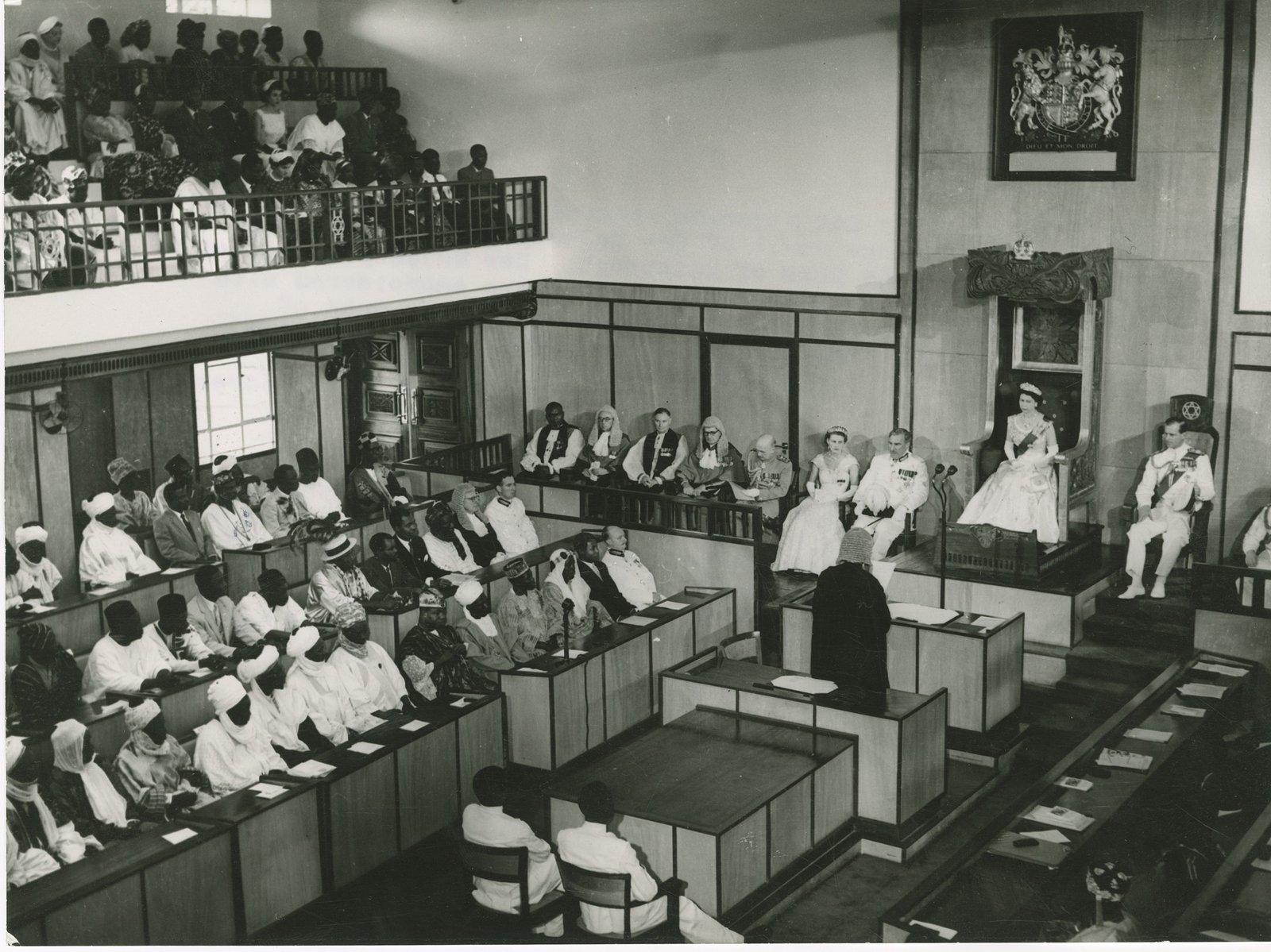 A crowded legislative chamber seen from the gallery: members in robes and caps on the benches at left, a public gallery above them, and Queen Elizabeth II on a carved throne beneath the royal arms at right, with the Duke of Edinburgh seated beside her.