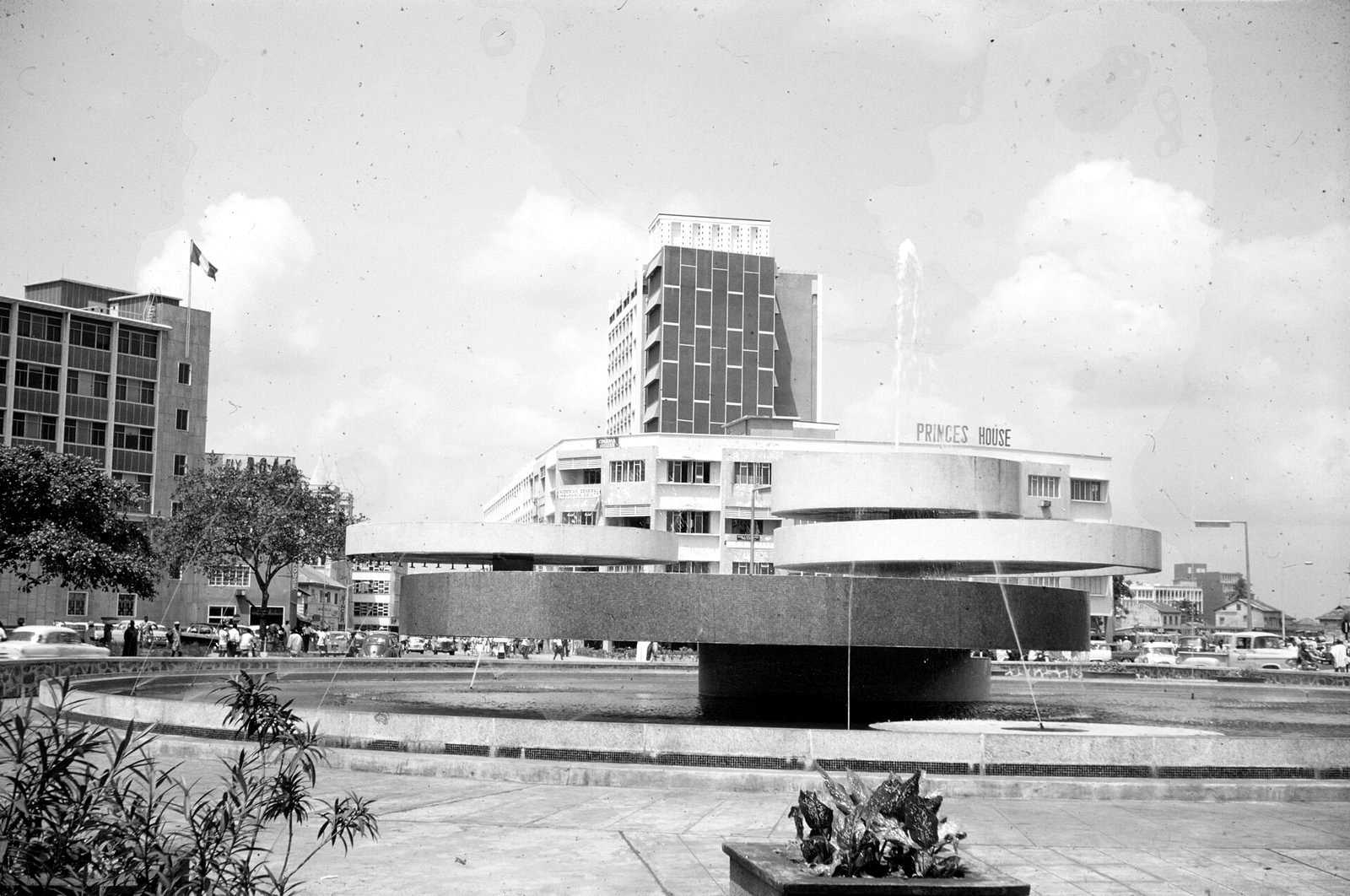 A round fountain with tiered concrete discs in a wide pool, in front of modern office buildings, one lettered Princes House, with a flag flying from a pole at left and traffic beyond.