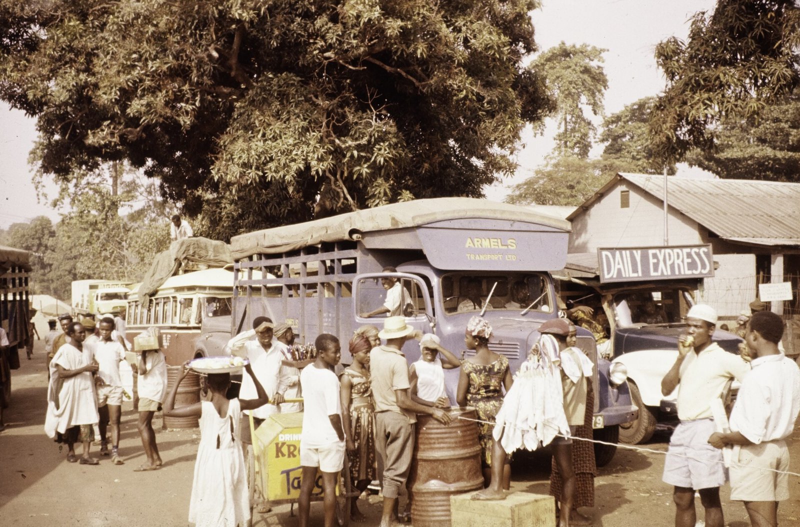 A crowd of men, women and children standing under a large tree beside a blue lorry marked Armels Transport Ltd, a loaded bus and a car under a sign reading Daily Express, with a drinks cart and a girl carrying a tray on her head.