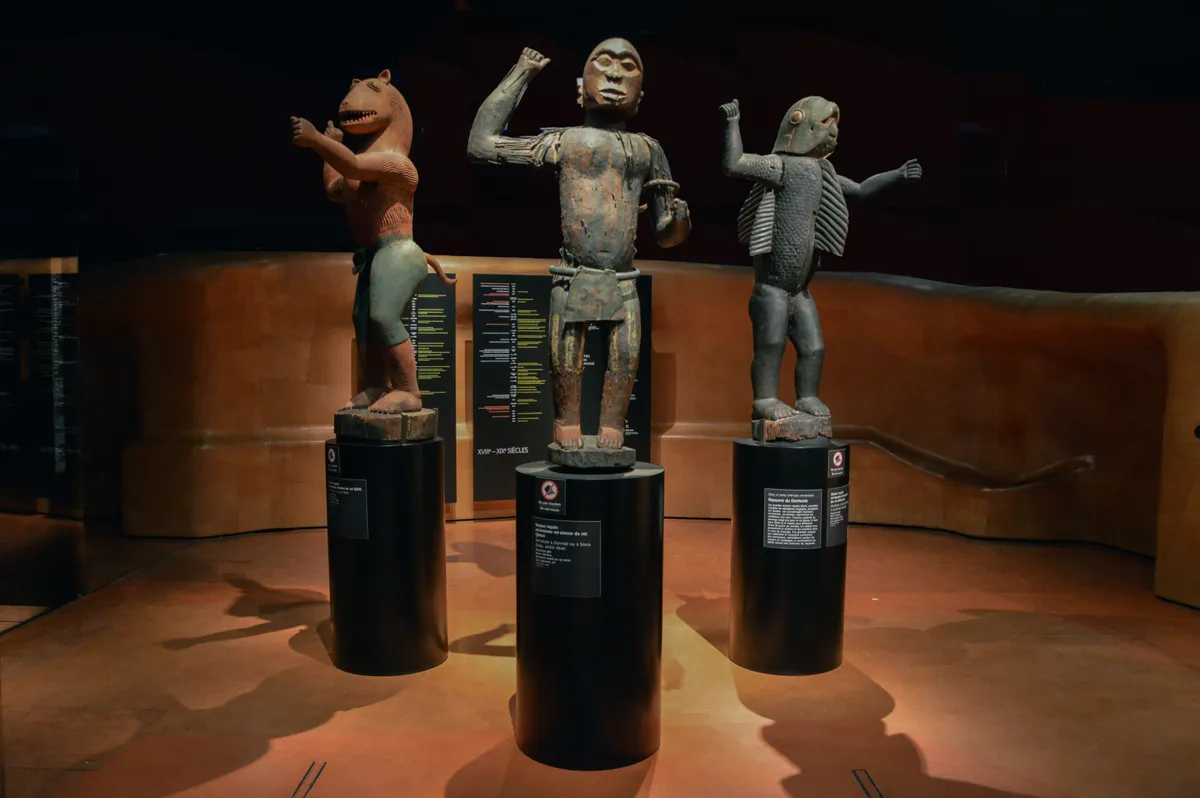 Three carved wooden royal figures on plinths in a dim gallery: one with a lion's head, one human with a raised arm, one bird-like.