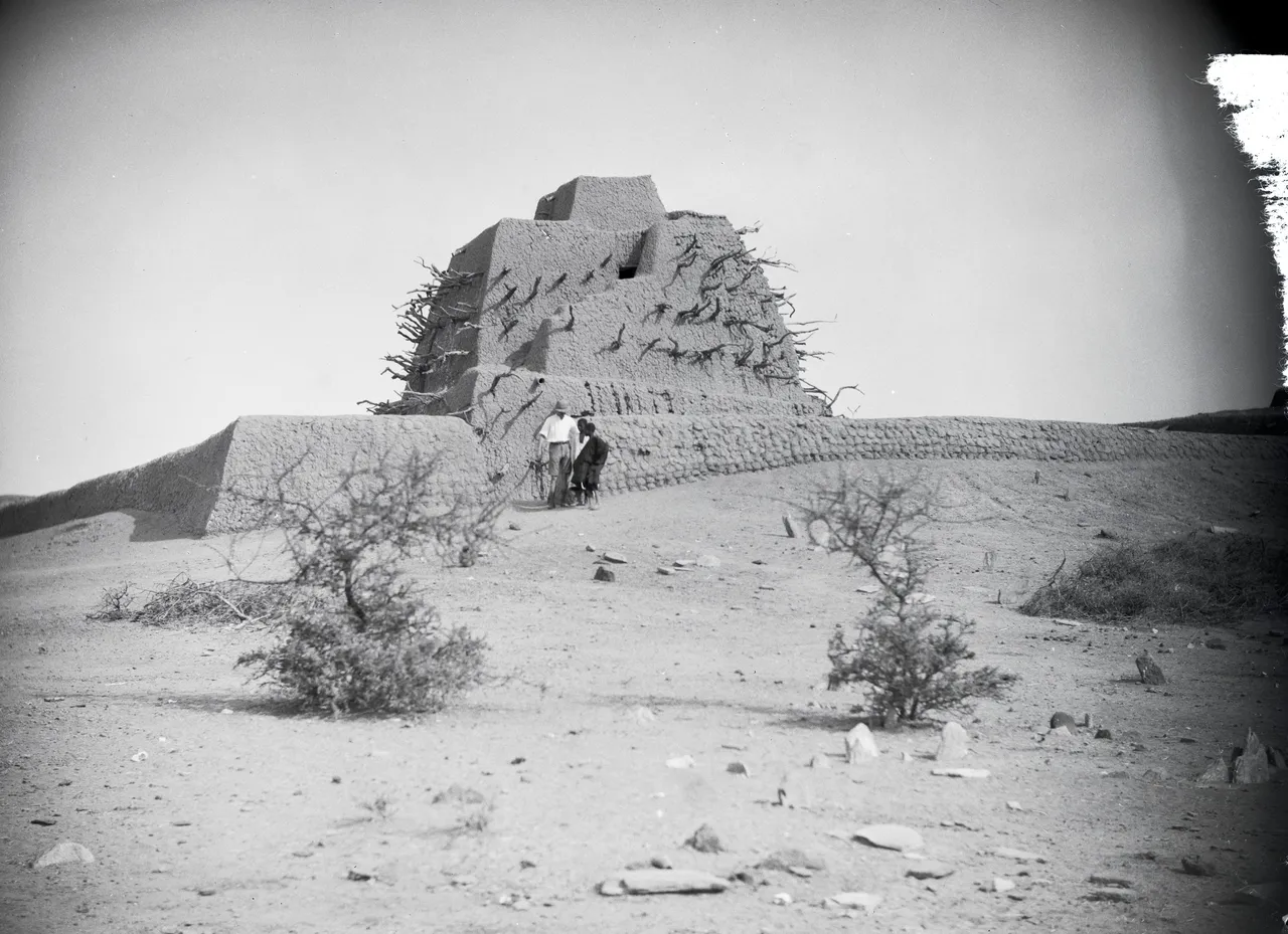 A black-and-white photograph of a stepped, pyramid-shaped mud-brick tomb bristling with wooden beams.