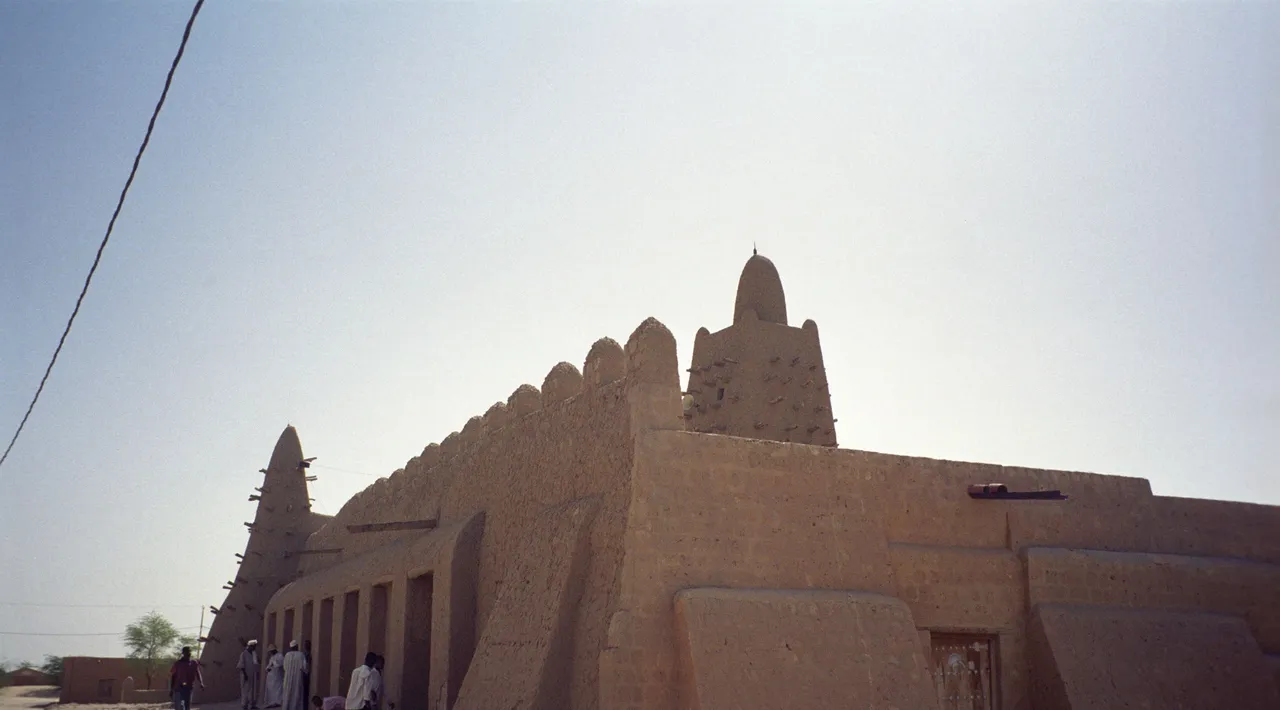 The long mud-brick wall and conical minaret of a mosque against a pale sky, with people walking by.