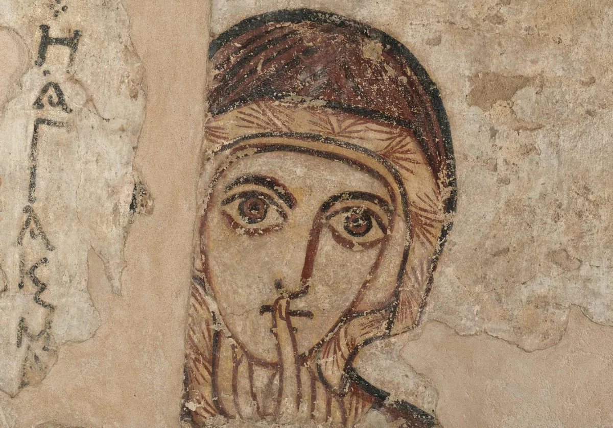 A painted woman's face with large dark eyes, one finger raised to her lips, on a worn ochre wall.