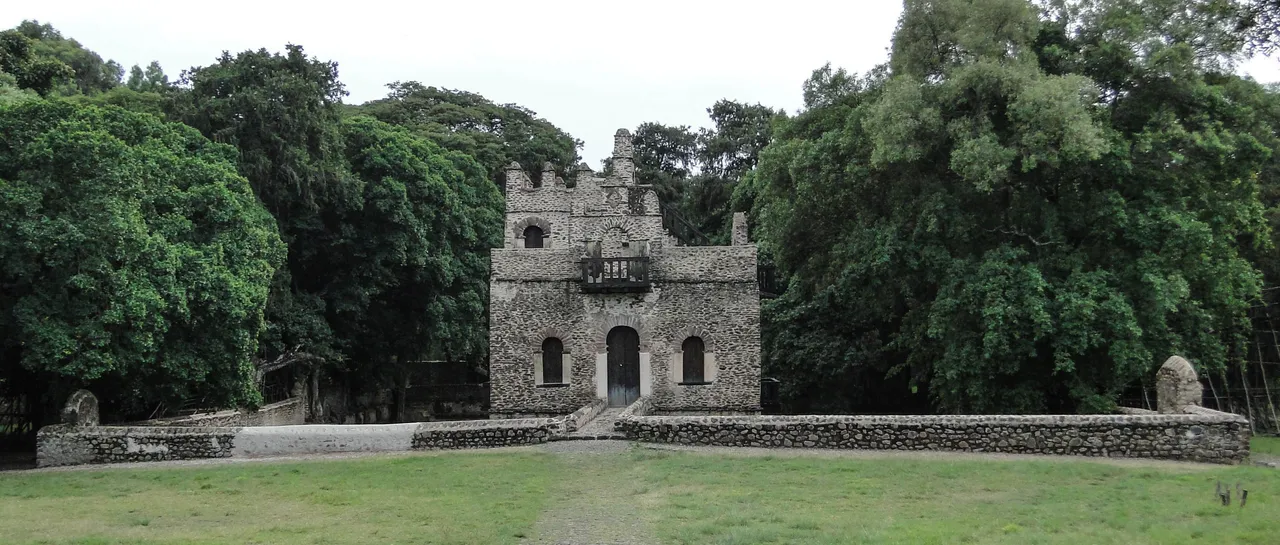 A small crenellated stone pavilion behind a low wall, framed by large green trees.