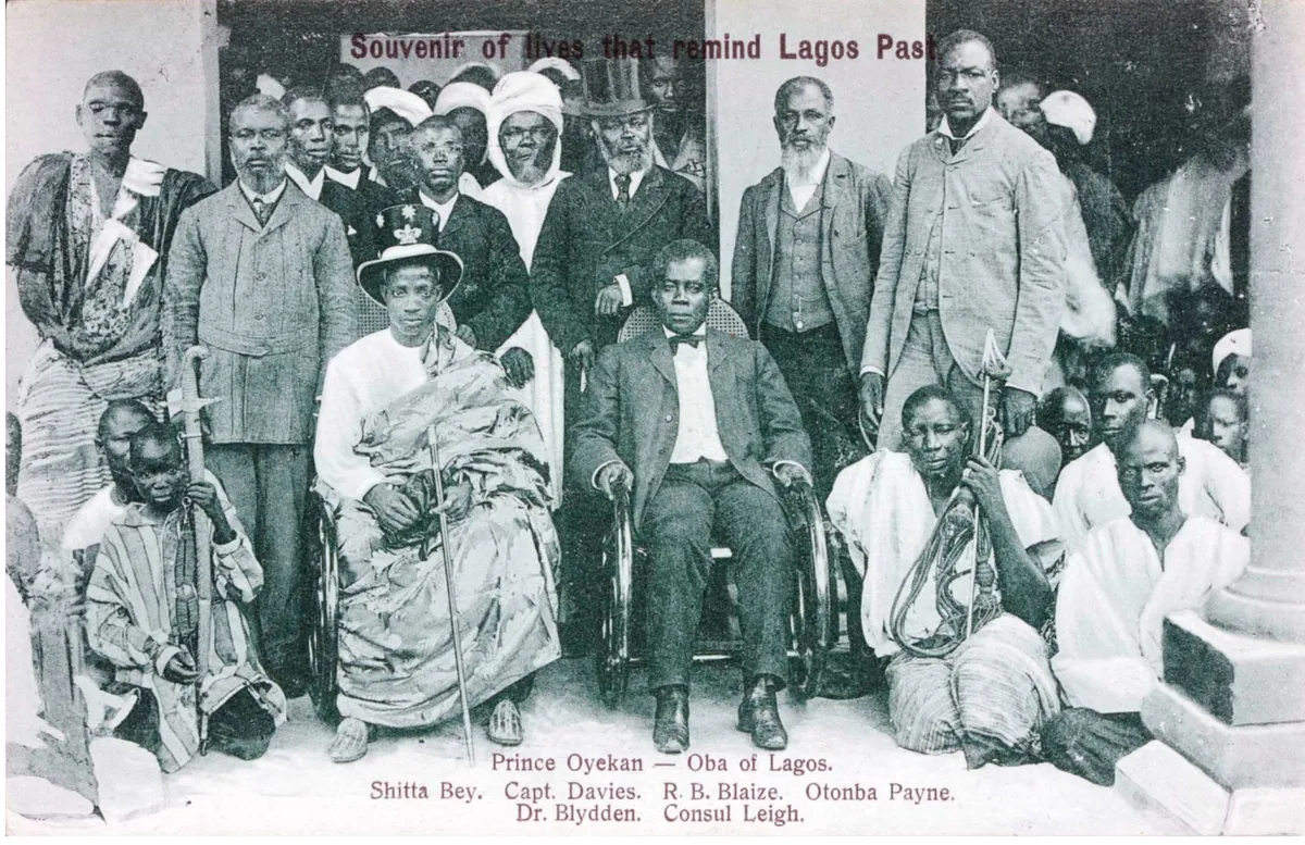 A black-and-white group photograph of men in robes and suits, two seated in front, standing before a building.