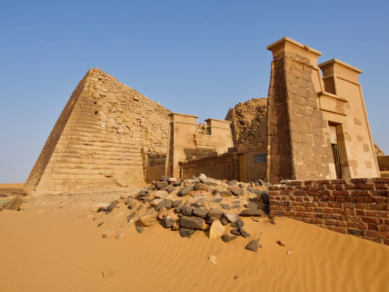 A steep sandstone pyramid with a small chapel at its base, on desert sand.