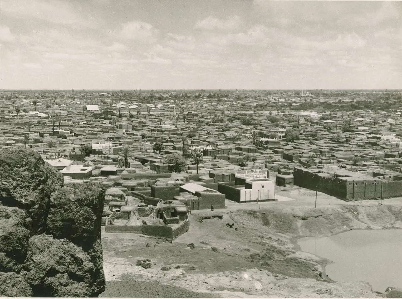 A dense city of flat-roofed mud-walled compounds seen from a height, with palms between the houses and a few whitewashed buildings.