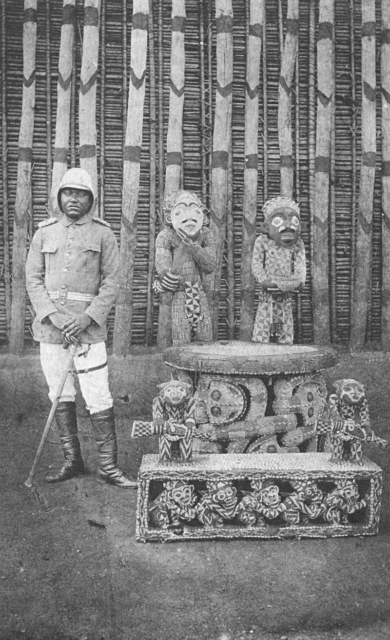 A black-and-white photograph of a man in a uniform and pith helmet standing beside a carved, beaded throne.