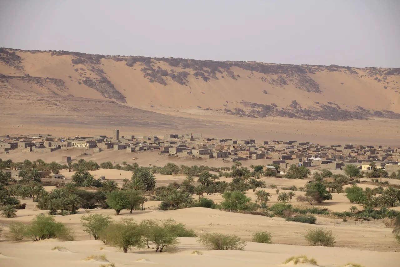 A low stone town among sand and scrub at the foot of a long desert escarpment.