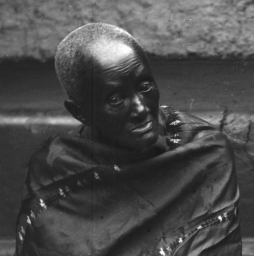 A black-and-white portrait of an elderly woman with short grey hair, wrapped in a dark cloth edged with small ornaments, seated against a plain wall.