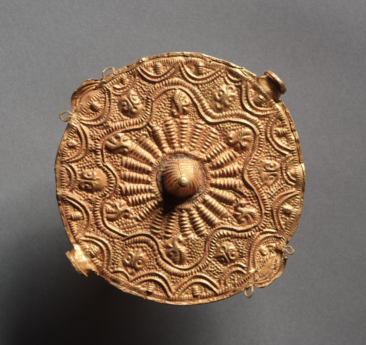 A round gold disc worked in repousse with radiating ribbed bands and small figures around a raised central boss, on a grey background.