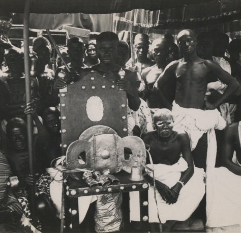A black-and-white photograph of a crowd of men around a chair on which rests a gold-covered stool hung with bells, with an elderly seated man beside it.