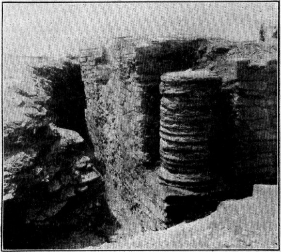 A grainy black-and-white photograph of a ruined stone building with a column built of stacked stone drums standing at the center.