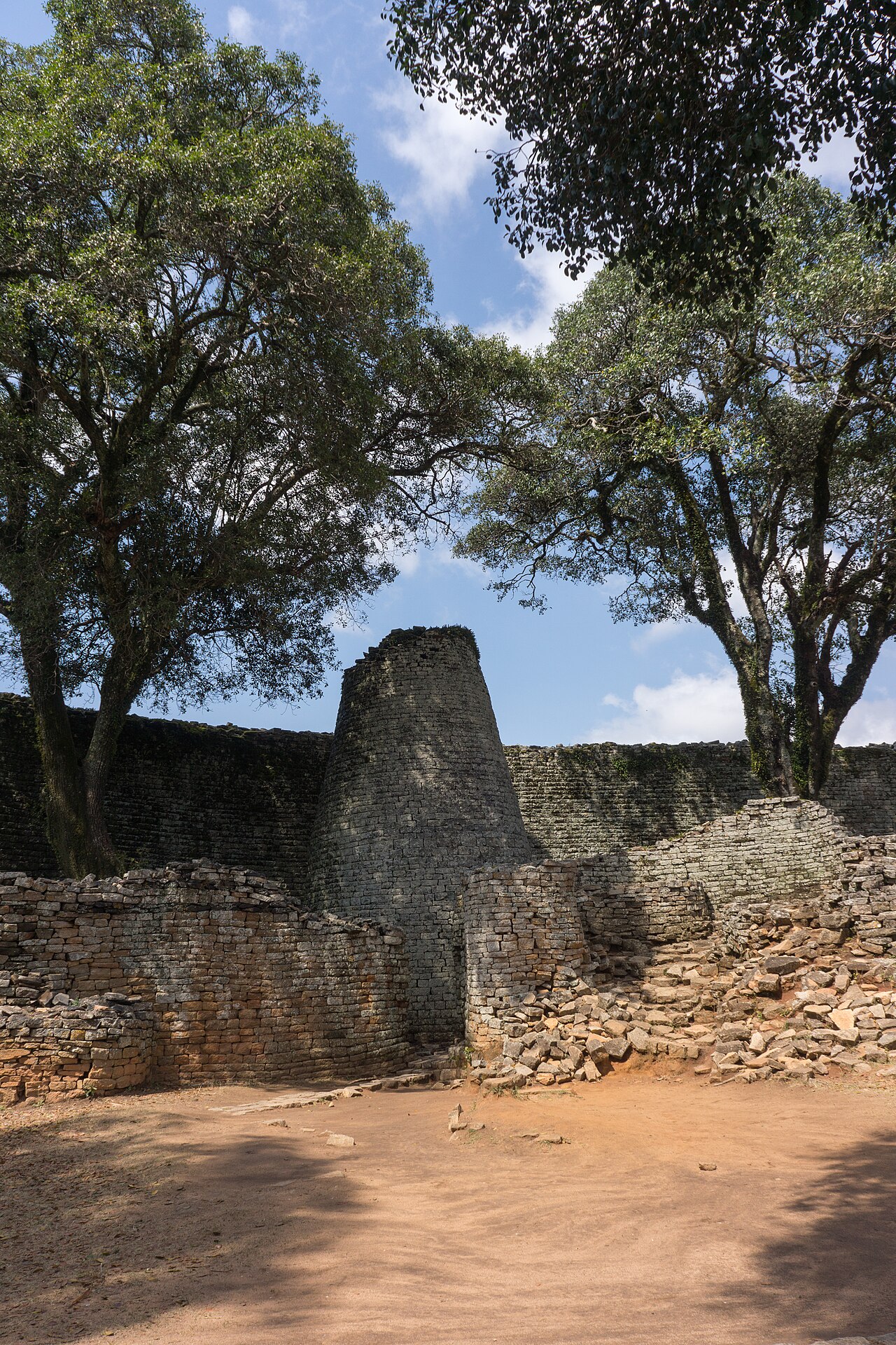 A solid, tapering tower of neatly coursed granite blocks standing inside a high stone wall, framed by trees.