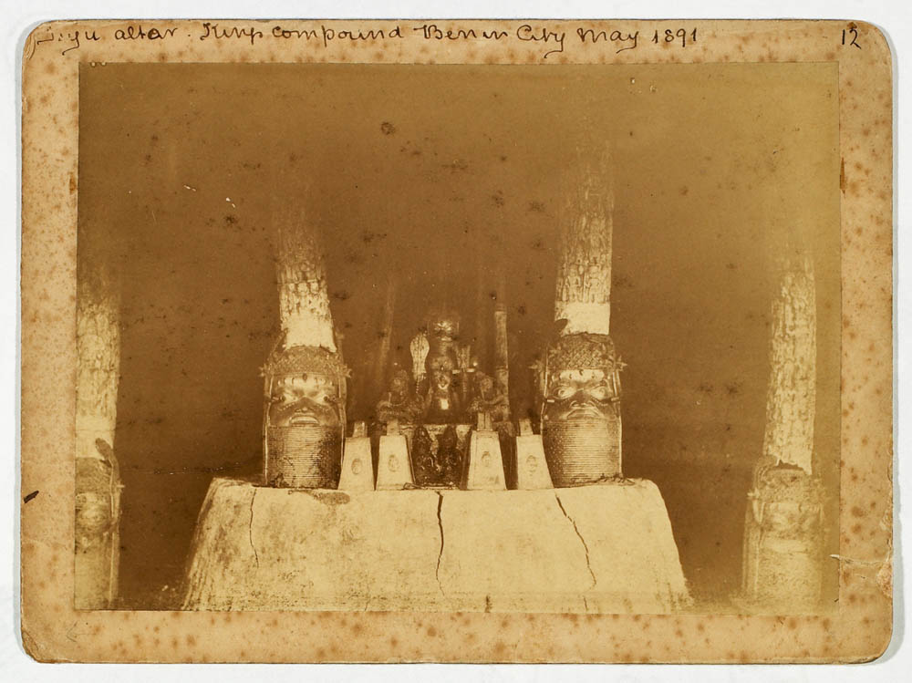 A faded sepia photograph of an altar in a dark room, with two cast brass heads, each topped by a tall carved ivory tusk, and smaller objects between them.
