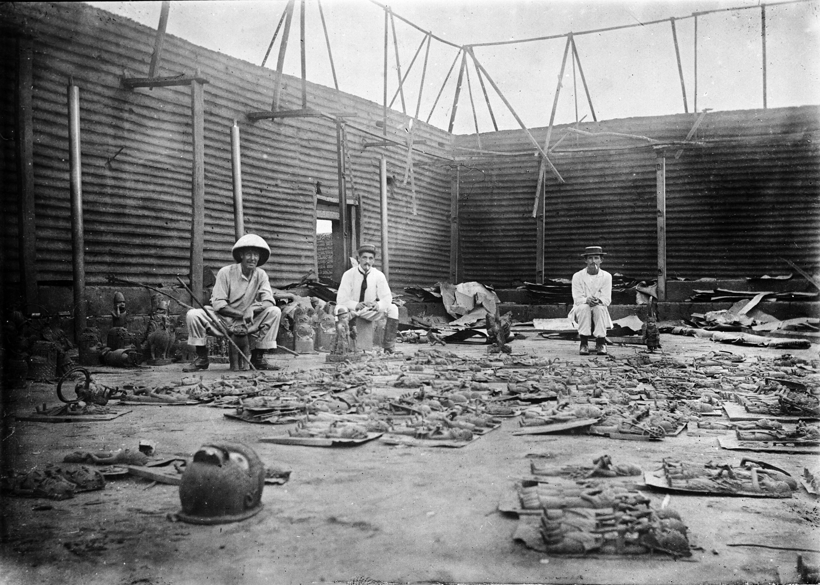 Three men in European clothes sit in a roofless courtyard with corrugated walls, surrounded by dozens of metal plaques and other objects laid out on the ground.