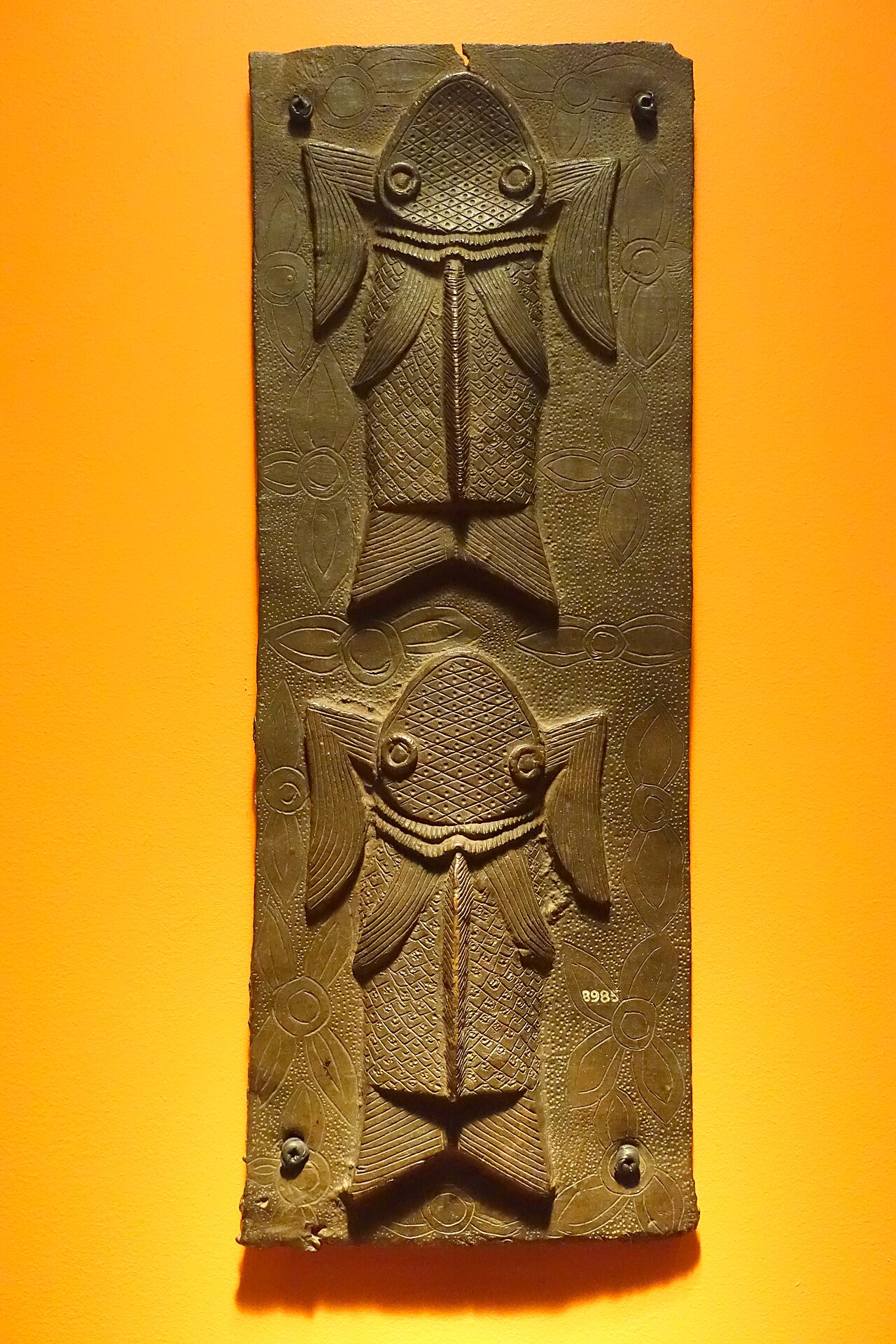 A long rectangular metal plaque with two stylized fish, one above the other, on a background of fine incised patterns, displayed against an orange wall.