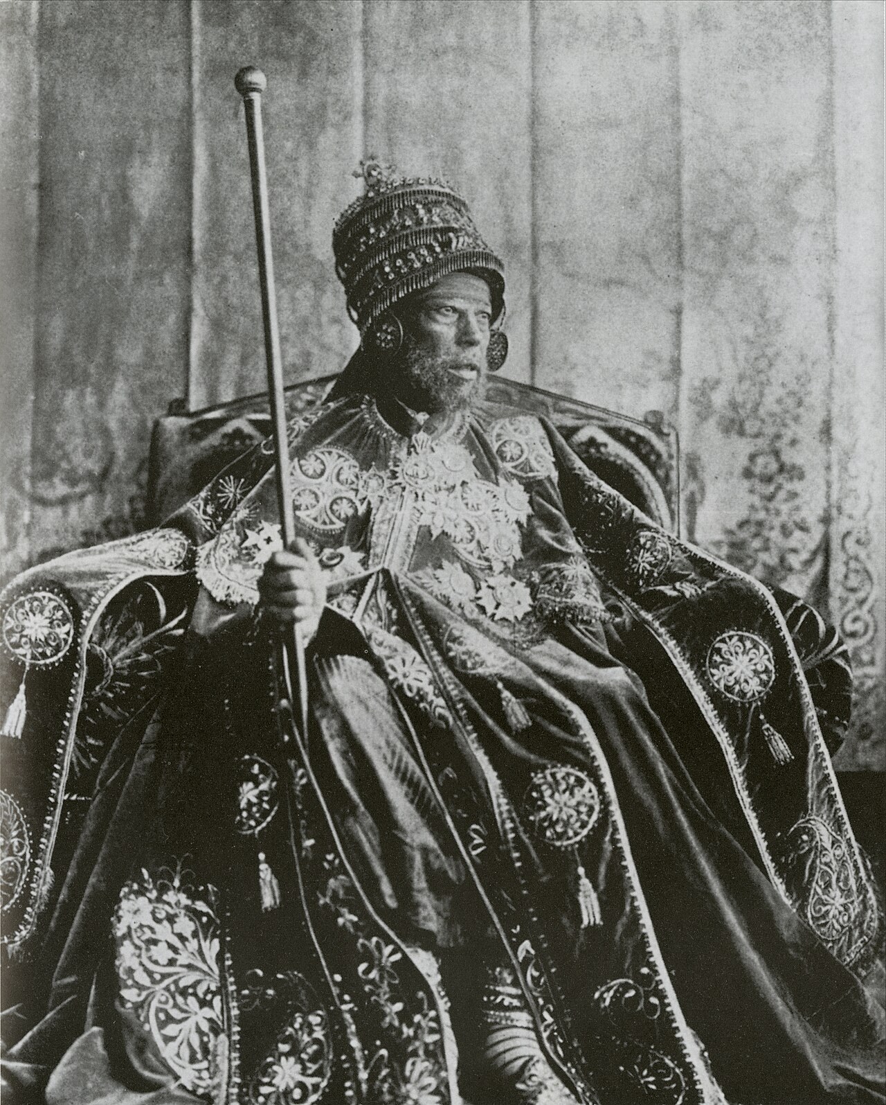 A black-and-white studio photograph of a bearded emperor seated on a throne in a tall crown and a heavily embroidered cloak with tassels, holding a long staff.