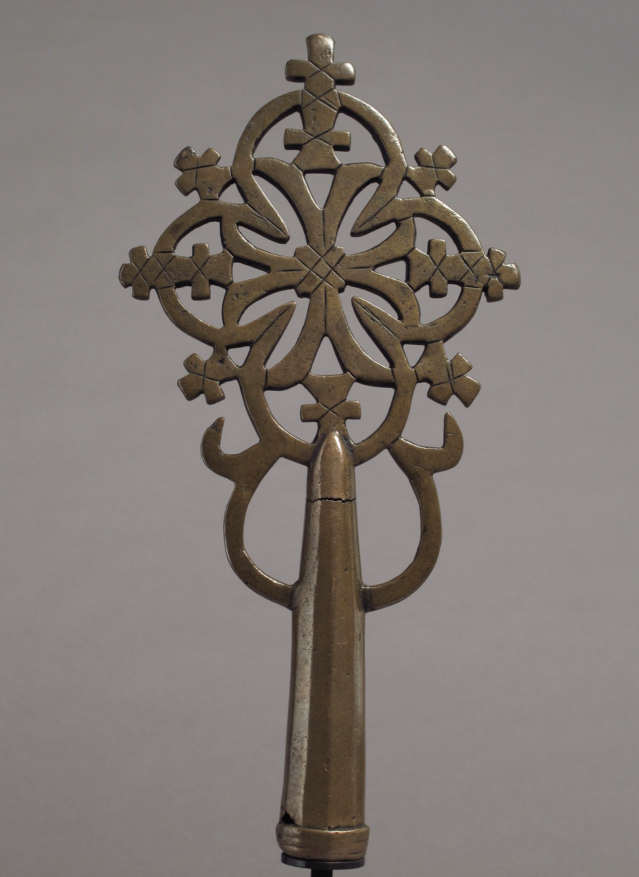 A bronze processional cross with an openwork head of interlacing loops and small crosses around a central cross, on a plain grey background.