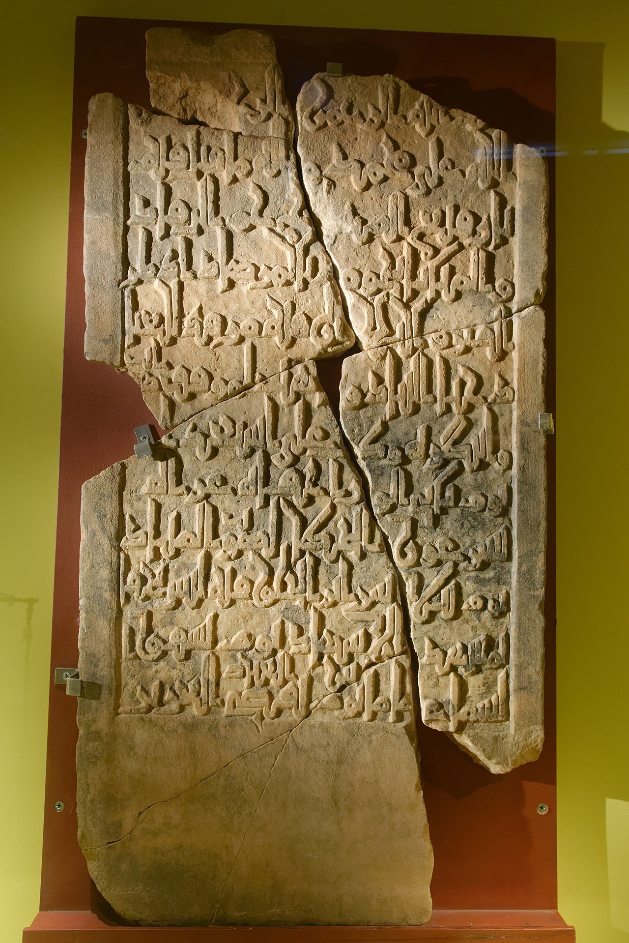 A tall, cracked marble slab carved with rows of angular Arabic script, displayed in a case.